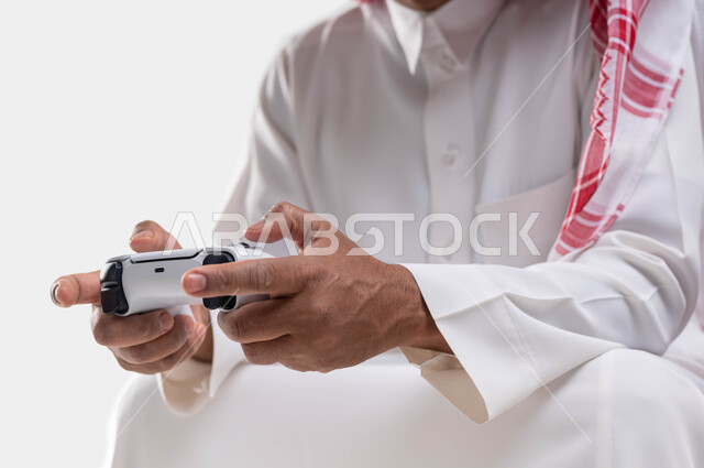 اجهزة وتقنيات الكترونية حديثة ترفيهيه, قضاء وقت ممتع ومسلي باللعب، التفاعل والاندماج مع ألعاب البلايستيشن، بورتريه مقرب لأيدي رجل عربي سعودي خليجي يرتدي الشماغ والثوب التقليدي يجلس على كرسي مريح يحمل بيده عصا التحكم يقوم باللعب في ألعاب الفيديو، خلفية بيضاء