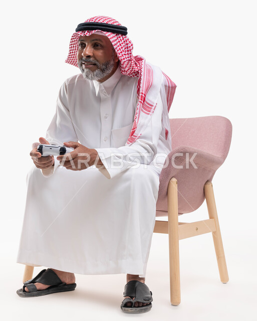 Modern electronic entertainment technologies, spending a fun and entertaining time playing, interacting and merging with PlayStation games, a portrait of an old Saudi Arabian Gulf man wearing a shemagh and traditional dress, sitting on a comfortable chair holding a joystick in his hand, playing video games, white background.