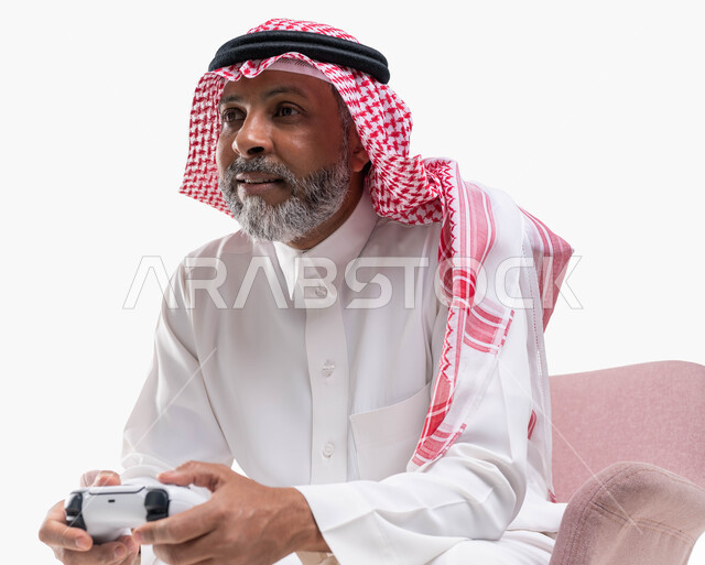 قضاء اوقات ممتعة ومسلية باللعب، التفاعل والاندماج مع ألعاب البلايستيشن، تقنيات الكترونية حديثة ترفيهيه, بورتريه مقرب لرجل عربي سعودي خليجي كبير بالسن يرتدي الشماغ والثوب التقليدي يجلس على كرسي مريح يحمل بيده عصا التحكم يقوم باللعب في ألعاب الفيديو، خلفية بيضاء