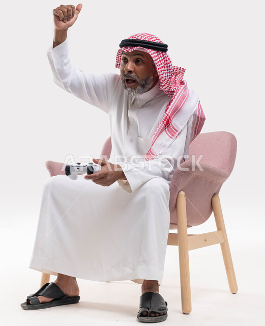 التفاعل والاندماج مع ألعاب البلايستيشن، ايماءات الانتصار والفوز، تقنيات الكترونية حديثة ترفيهيه, بورتريه لرجل عربي سعودي خليجي كبير بالسن يرتدي الشماغ والثوب التقليدي يجلس على كرسي مريح يحمل بيده عصا التحكم يقوم باللعب في ألعاب الفيديو، خلفية بيضاء