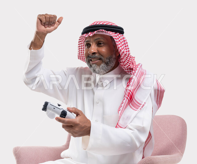 ايماءات الانتصار والفوز، التفاعل والاندماج مع ألعاب البلايستيشن، تقنيات الكترونية حديثة ترفيهيه, بورتريه مقرب لرجل عربي سعودي خليجي كبير بالسن يرتدي الشماغ والثوب التقليدي يجلس على كرسي مريح يحمل بيده عصا التحكم يقوم باللعب في ألعاب الفيديو، خلفية بيضاء
