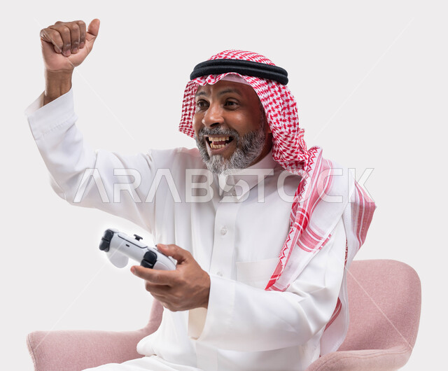 تعابير الفوز والانتصار، التفاعل والاندماج مع ألعاب البلايستيشن، تقنيات الكترونية حديثة ترفيهيه, بورتريه مقرب لرجل عربي سعودي خليجي كبير بالسن يرتدي الشماغ والثوب التقليدي يجلس على كرسي مريح يحمل بيده عصا التحكم يقوم باللعب في ألعاب الفيديو، خلفية بيضاء