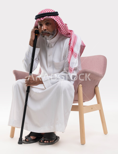 النظر الى شيء ما بإيماءات التفكير والشرود في الماضي وأيام الشباب، مفهوم الجلوس والراحة، بورتريه لرجل عربي خليجي سعودي كبير بالسن يرتدي الشماغ والثوب التقليدي يجلس على كرسي مريح يستند على عكازه، خلفية بيضاء