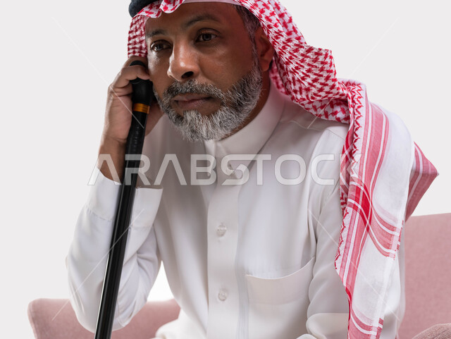Feeling lethargic and bored, the concept of sitting and resting, looking at something with gestures of thought and distraction, a close-up portrait of an old Saudi Gulf Arab man wearing a shemagh and traditional dress, leaning on his cane, white background.