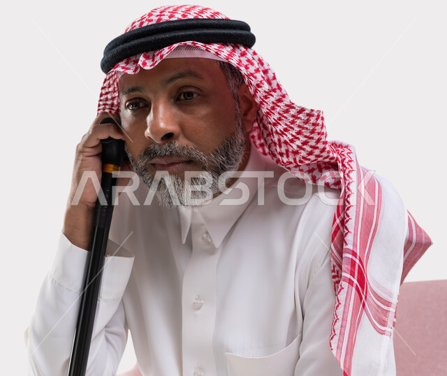 The concept of sitting and resting, looking at something with gestures of thought and distraction, a close-up portrait of an old Saudi Gulf Arab man wearing a shemagh and traditional thobe, leaning on his cane, feeling tired and lethargic, white background.