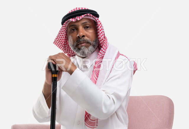 إيماءات التحديق والتفكير والتأمل، أدوات للمساعدة في المشي، بورتريه لرجل كبير السن عربي خليجي سعودي يرتدي الشماغ والثوب التقليدي ينظر إلى الكاميرا يمسك بيده العكازة، الأناقة والاهتمام بالمظهر الخارجي، خلفية بيضاء