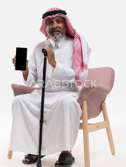 استخدام الهاتف المحمول بشاشة سوداء فارغة، عرض برامج وتطبيقات الجوال، تصفح مواقع التواصل الاجتماعي، بورتريه لرجل كبير السن عربي خليجي سعودي يرتدي الشماغ والثوب التقليدي ينظر إلى الكاميرا يمسك بيده العكازة، خلفية بيضاء