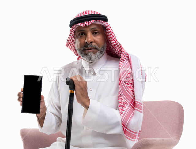 عرض برامج وتطبيقات الجوال، استخدام الهاتف المحمول بشاشة سوداء فارغة، تصفح مواقع التواصل الاجتماعي، بورتريه لرجل كبير السن عربي خليجي سعودي يرتدي الشماغ والثوب التقليدي ينظر إلى الكاميرا يمسك بيده العكازة، خلفية بيضاء