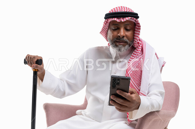 استخدام التقنيات الحديثة والمتطورة، الدردشة مع الأصدقاء عبر برامج وتطبيقات الجوال، تصفح مواقع التواصل الاجتماعي، بورتريه لرجل كبير السن عربي خليجي سعودي يرتدي الشماغ والثوب التقليدي يستخدم الهاتف المحمول يمسك بيده العكازة، خلفية بيضاء