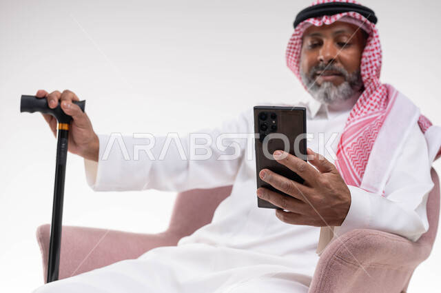 التواصل مع الأصدقاء عبر برامج وتطبيقات الجوال، تصفح مواقع التواصل الاجتماعي، بورتريه لرجل كبير السن عربي خليجي سعودي يرتدي الشماغ والثوب التقليدي يستخدم الهاتف المحمول يمسك بيده العكازة، استخدام تقنيات حديثة ومتطورة، خلفية بيضاء
