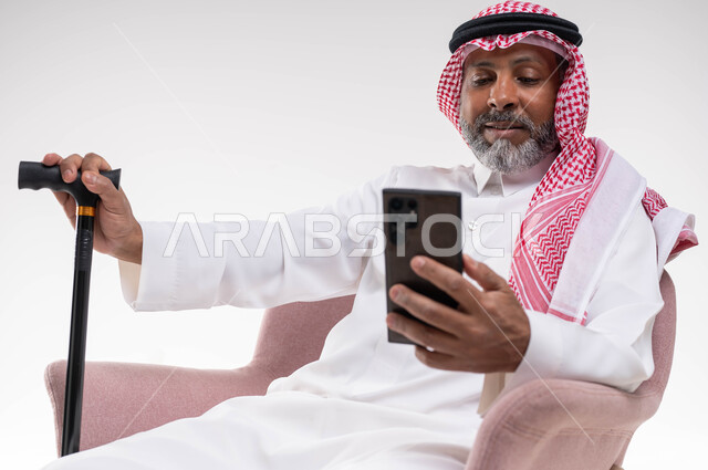 استخدام التقنيات الحديثة والمتطورة، تصفح مواقع التواصل الاجتماعي، بورتريه لرجل كبير السن عربي خليجي سعودي يرتدي الشماغ والثوب التقليدي يحمل الهاتف المحمول يمسك بيده العكازة، التواصل مع الأصدقاء عبر برامج وتطبيقات الجوال، خلفية بيضاء