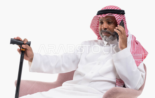 إجراء مكالمات صوتية بإيماءات التفكير، دردشات ومحادثات عبر الانترنت، بورتريه لرجل كبير السن عربي خليجي سعودي يرتدي الشماغ والثوب التقليدي يستخدم الهاتف المحمول يمسك بيده العكازة، التواصل مع الأصدقاء عبر برامج التواصل الاجتماعي، خلفية بيضاء.