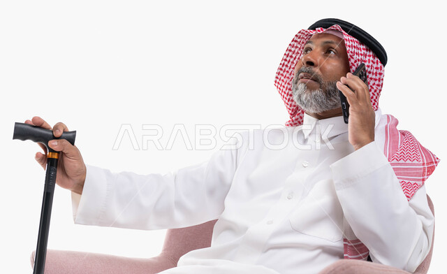 التواصل مع الأصدقاء عبر برامج التواصل الاجتماعي، دردشات ومحادثات عبر الانترنت، بورتريه لرجل كبير السن عربي خليجي سعودي يرتدي الشماغ والثوب التقليدي يستخدم الهاتف المحمول يمسك بيده العكازة، إجراء مكالمات صوتية بإيماءات التفكير، خلفية بيضاء.