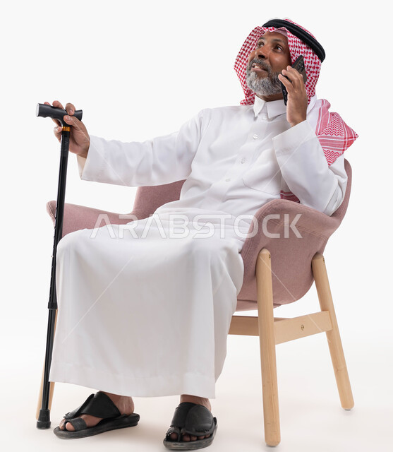 دردشات ومحادثات عبر الانترنت، بورتريه لرجل كبير السن عربي خليجي سعودي يرتدي الشماغ والثوب التقليدي يستخدم الهاتف المحمول يجلس على كرسي مريح يمسك بيده العكازة، إجراء مكالمات صوتية بإيماءات السعادة، التواصل مع الأصدقاء عبر برامج التواصل الاجتماعي، خلفية بيضاء.