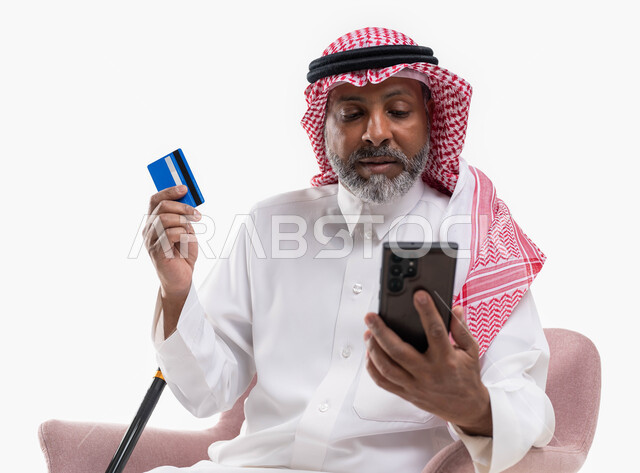 Using mobile applications to pay and purchase, advanced modern technologies, paying bills by electronic card, a close-up portrait of an elderly Saudi Gulf Arab man wearing a shemagh and traditional dress, holding a mobile phone and bank credit card in his hand, white background