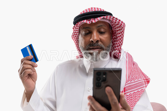 التقنيات الحديثة المتطورة، تسديد الفواتير بالبطاقة الإلكترونية، الشراء والدفع من خلال برامج وتطبيقات الجوال، بورتريه مقرب لرجل كبير السن  عربي خليجي سعودي يرتدي الشماغ والزي التقليدي يمسك بيده الهاتف المحمول وبطاقة الإئتمان البنكية، خلفية بيضاء