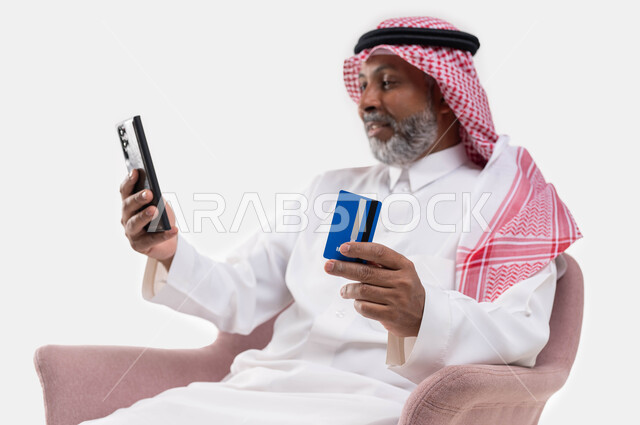 إيماءات السعادة والفرح، الشراء والدفع من خلال برامج وتطبيقات الجوال، بورتريه مقرب لرجل كبير السن عربي خليجي سعودي مبتسم يرتدي الشماغ والزي التقليدي ويمسك بيده الهاتف المحمول وبطاقة الإئتمان البنكية، إمكانية تسديد الفواتير بالبطاقة الإلكترونية، خلفية بيضاء