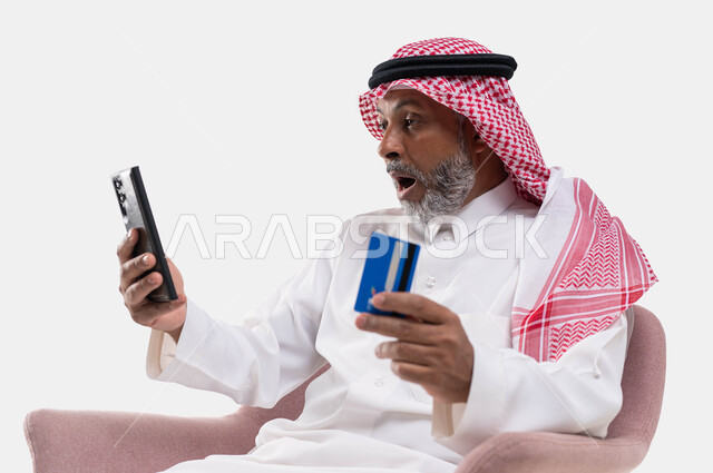 Gestures of astonishment and astonishment, payment through mobile programs and applications, a close-up portrait of an elderly, smiling Saudi Gulf Arab man wearing a shemagh and traditional dress, holding a mobile phone and bank credit card in his hand, the ability to pay bills by electronic card, white background