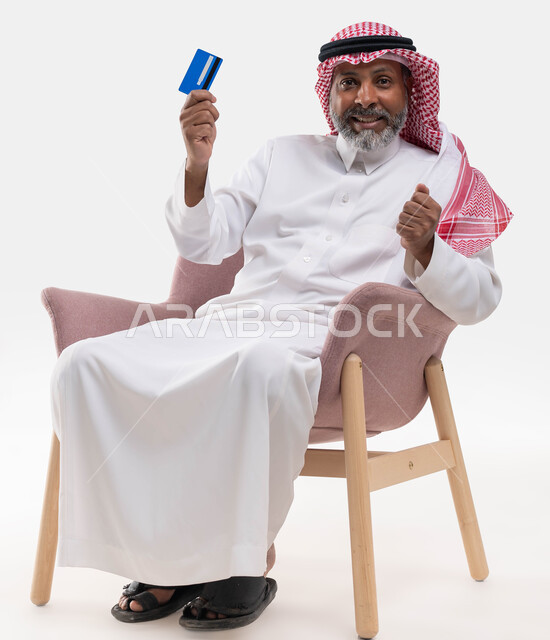 إيماءات السعادة والفرح، بورتريه مقرب لرجل كبير السن عربي خليجي سعودي مبتسم يرتدي الشماغ والزي التقليدي يجلس على كرسي مريح ينظر إلى الكاميرا يمسك بيده بطاقة الإئتمان البنكية، إمكانية تسديد الفواتير بالبطاقة الإلكترونية، خلفية بيضاء