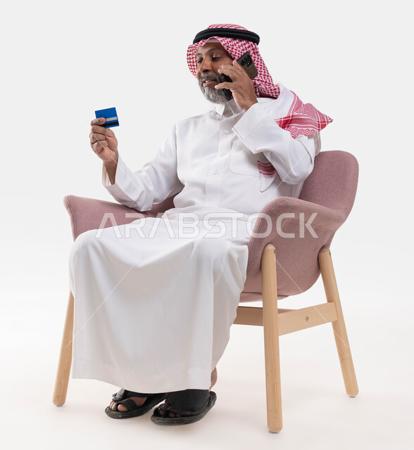 Sitting in a comfortable chair and making voice calls, paying through mobile applications, a close-up portrait of an elderly, smiling Saudi Arabian Gulf man wearing a shemagh and traditional dress, holding a mobile phone and bank credit card in his hand, the ability to pay bills by electronic card, white background