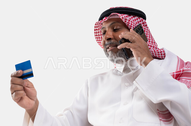 Payment and purchase through mobile programs and applications, a close-up portrait of an elderly, smiling Saudi Arabian Gulf man wearing a shemagh and traditional dress, holding a mobile phone and a bank credit card in his hand, the ability to pay bills by electronic card, making a voice call, white backgroun