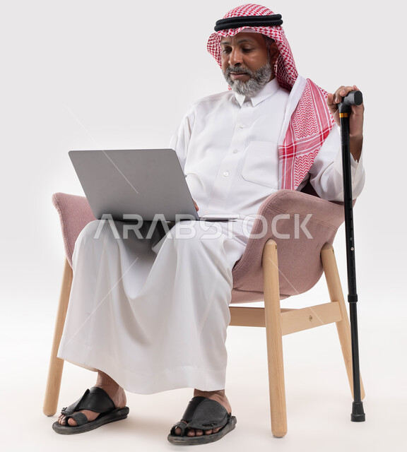 إجراء مكالمة فيديو وبث مباشر عبر جهاز الكمبيوتر، تصفح المواقع الالكترونية، العمل عن بعد باستخدام الحاسوب المحمول، بورتريه مقرب لرجل شايب عربي سعودي خليجي يرتدي الشماغ والزي التقليدي يجلس على كرسي مريح يمسك العكاز للمساعدة في النهوض والمشي، خلفية بيضاء