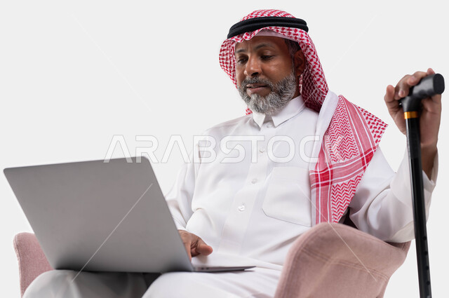 Working remotely using a laptop, live broadcasting and making a video call via the computer, a close-up portrait of an elderly Saudi Arabian Gulf man wearing a shemagh and traditional dress, holding a crutch to help with walking and getting up, browsing websites, white backgroun