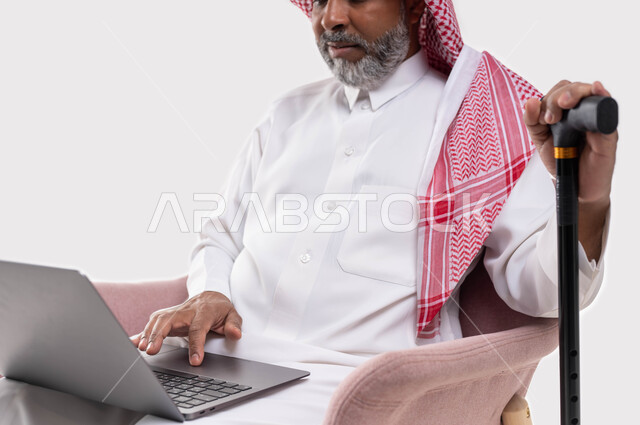 بث مباشر واجراء مكالمة فيديو عبر جهاز الكمبيوتر، العمل عن بعد باستخدام الحاسوب المحمول، بورتريه مقرب لرجل شايب عربي سعودي خليجي مبتسم يرتدي الشماغ والزي التقليدي يمسك العكاز للمساعدة في المشي والنهوض، تصفح المواقع الالكترونية، خلفية بيضاء