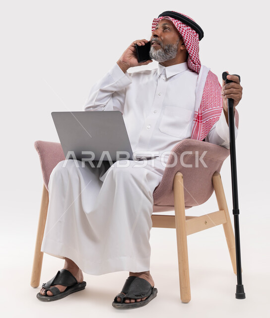 Tools to help with walking, achieving strategies and goals, a close-up portrait of an elderly Saudi Arabian Gulf man wearing traditional clothing and a shemagh, holding a crutch, making a call on a mobile phone, working on a laptop, completing tasks online through a computer, white background