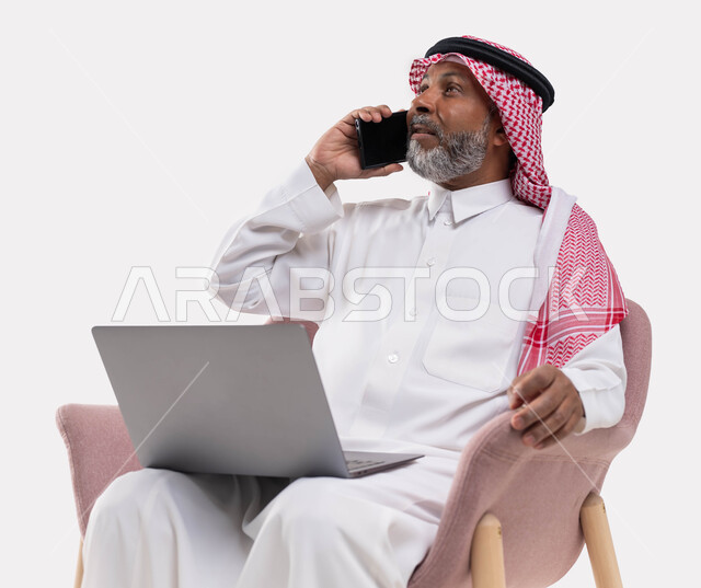 تحقيق الاستراتيجيات والأهداف، استخدام التقنيات الحديثة في العمل عن بعد، بورتريه مقرب  لرجل كبير السن عربي خليجي سعودي يرتدي الزي التقليدي والشماغ يجري مكالمة عبر الهاتف المحمول يعمل عبر اللابتوب، إنجاز المهام اونلاين من خلال جهاز الحاسوب، خلفية بيضاء .