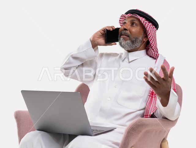 إنجاز المهام اونلاين من خلال جهاز الحاسوب، استخدام التقنيات الحديثة في العمل عن بعد، بورتريه مقرب  لرجل كبير السن عربي خليجي سعودي يرتدي الزي التقليدي والشماغ يجري مكالمة عبر الهاتف المحمول يعمل عبر اللابتوب، تحقيق الاستراتيجيات والأهداف، خلفية بيضاء .