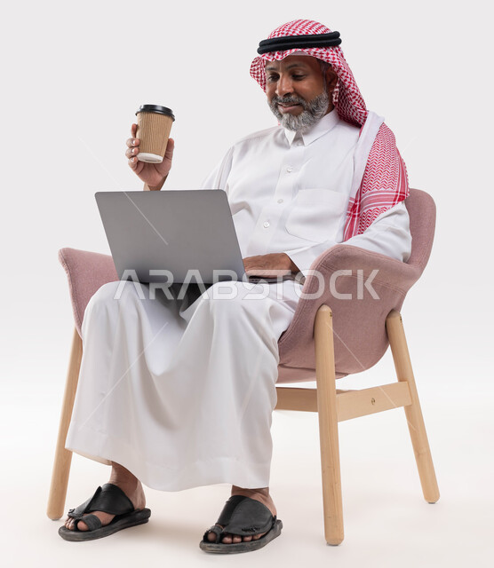 استخدم الحاسب المحمول لآداء مهام العمل, احتساء المشروبات الشتوية الساخنه، الاستمتاع بتناول القهوة العربية، بورتريه لرجل شايب عربي خليجي سعودي مبتسم يرتدي الشماغ والزي التقليدي يحمل بيده كوب من الورق المقوى يجلس على كرسي مريح، خلفية بيضاء