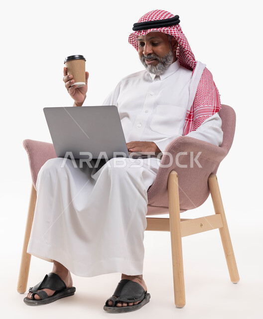 Sipping hot winter drinks, enjoying Arabic coffee, sitting in a comfortable chair and working on the laptop, portrait of an elderly Saudi Gulf Arab man, smiling, wearing a shemagh and traditional dress, holding a cardboard cup in his hand, using a laptop, white backgroun