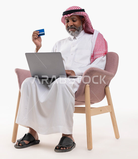 إمكانية تسديد الفواتير بالبطاقة الإلكترونية، الشراء والدفع من خلال برامج وتطبيقات اللابتوب، بورتريه لرجل شايب عربي خليجي سعودي مبتسم يرتدي الشماغ والثوب التقليدي يجلس على كرسي مريح يمسك بيده بطاقة الإئتمان البنكية يستخدم جهاز الحاسب المحمول،  خلفية بيضاء