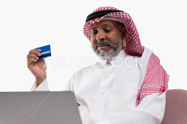 الشراء والدفع من خلال برامج وتطبيقات اللابتوب، بورتريه لرجل شايب عربي خليجي سعودي مبتسم يرتدي الشماغ والثوب التقليدي يمسك بيده بطاقة الإئتمان البنكية يستخدم جهاز الحاسب المحمول، إمكانية تسديد الفواتير بالبطاقة الإلكترونية، خلفية بيضاء