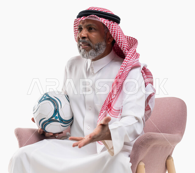 متابعة ومشاهدة مباراة كرة القدم بإيماءات الانزعاج لخسارة الفريق، التفاعل مع المنتخب، بورتريه لرجل كبير السن عربي خليجي سعودي يرتدي الشماغ والثوب التقليدي يجلس على كرسي مريح يحمل الكرة، خلفية بيضاء