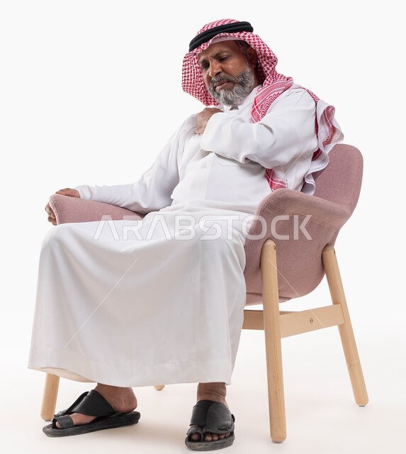 الجلوس على الكرسي والشعور بالتعب والألم الشديد, بورتريه لرجل كبير بالسن عربي خليجي سعودي مريض يرتدي الثوب التقليدي والشماغ يضع يديه على كتفه ويشعر بالوجع, إيماءات الانزعاج والحاجة إلى العلاج والدواء, خلفية بيضاء