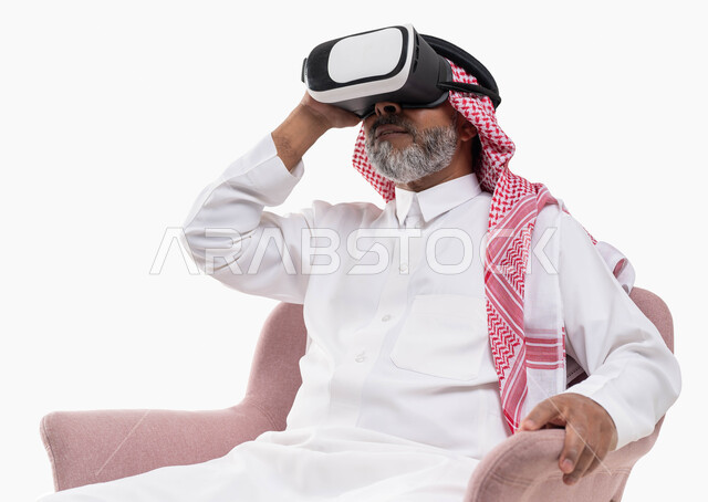 قضاء وقت ممتع بمشاهدة الأفلام عبر نظارة الواقع الافتراضي, التفاعل والاندماج ومحاكاة العالم الخيالي, بورتريه لرجل كبير بالسن عربي خليجي سعودي يرتدي الثوب التقليدي والنظارة ثلاثية الابعاد يجلس على كرسي مريح, خلفية بيضاء