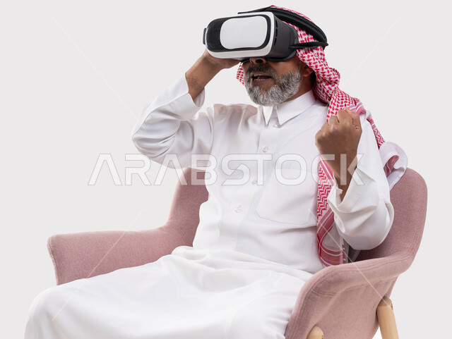 الاستمتاع بمشاهدة الأفلام عبر نظارة الواقع الافتراضي, التفاعل والاندماج ومحاكاة العالم الخيالي, بورتريه لرجل كبير بالسن عربي خليجي سعودي يرتدي الثوب التقليدي والنظارة ثلاثية الابعاد يجلس على كرسي مريح ويرفع يده بإيماءات الإعجاب, خلفية بيضاء
