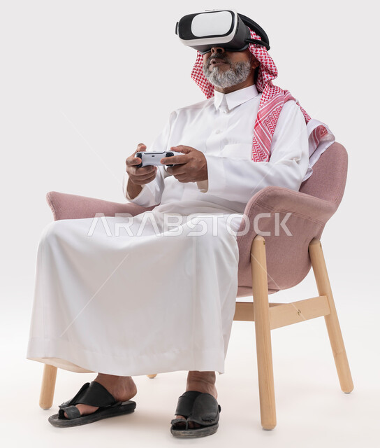 قضاء وقت ممتع بمشاهدة الأفلام عبر نظارة الواقع الافتراضي, التفاعل والاندماج ومحاكاة العالم الخيالي, بورتريه لرجل كبير بالسن عربي خليجي سعودي يرتدي الثوب التقليدي والنظارة ثلاثية الابعاد يجلس على كرسي مريح ويمسك عصا التحكم, خلفية بيضاء