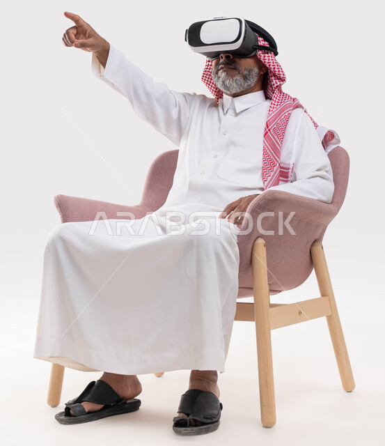 الاندماج ومحاكاة العالم الخيالي والاشارة إلى شيء ما, مشاهدة الأفلام عبر نظارة الواقع الافتراضي في آر, بورتريه لرجل كبير بالسن عربي خليجي سعودي يرتدي الثوب التقليدي والنظارة ثلاثية الابعاد يجلس على كرسي مريح ويرفع اصبعه للأعلى, خلفية بيضاء