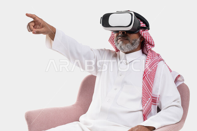 الإشارة إلى شيء ما باندماج ومحاكاة العالم الخيالي, مشاهدة الأفلام عبر نظارة الواقع الافتراضي في آر, بورتريه مقرب لرجل كبير بالسن عربي خليجي سعودي يرتدي الثوب التقليدي والنظارة ثلاثية الابعاد يرفع يديه للأعلى, خلفية بيضاء
