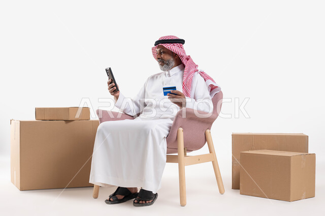 الدفع والشراء عن بعد عبر الاونلاين, استخدام بطاقة الإئتمان في التسوق الالكتروني، بورتريه مقرب لرجل كبير بالسن عربي سعودي خليجي يرتدي الشماغ والثوب التقليدي يجلس على كرسي يمسك البطاقة البنكية ويقوم بتصفح تطبيقات المصارف والبنوك عبر الجوال، خلفية بيضاء
