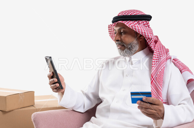 الشراء والدفع عن بعد عبر الاونلاين, استخدام بطاقة الإئتمان في التسوق الالكتروني، بورتريه مقرب لرجل  كبير بالسن عربي سعودي خليجي يرتدي الشماغ والثوب التقليدي يجلس على كرسي يمسك البطاقة البنكية ويقوم بتصفح تطبيقات المصارف والبنوك عبر الجوال، خلفية بيضاء
