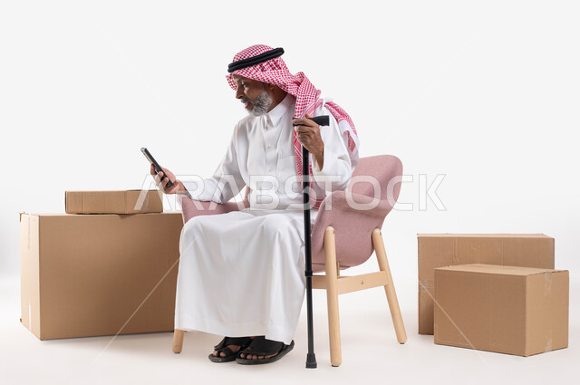 التواصل مع الأصدقاء عبر برامج وتطبيقات الهاتف المحمول، تصفح مواقع السوشال ميديا، بورتريه لرجل كبير السن عربي خليجي سعودي يرتدي الشماغ والثوب التقليدي يمسك بيده العكازة، استخدام الأجهزة والتقنيات الحديثة والمتطورة، خلفية بيضاء