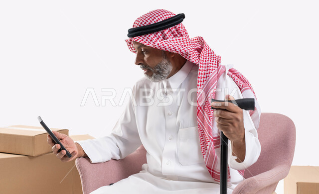 Using modern and advanced devices and technologies, communicating with friends via mobile phone programs and applications, browsing social media sites, a close-up portrait of an elderly Saudi Gulf Arab man wearing a shemagh and traditional thobe, holding a crutch in his hand, a white background.