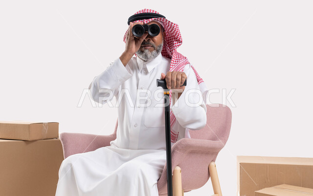 رؤية الأشياء البعيدة, الجلوس على الكرسي بإيماءات التركيز والاستكشاف, بورتريه لرجل كبير بالسن عربي خليجي سعودي يرتدي الثوب والتقليدي والشماغ يمسك بيده العكاز ويقوم بالنظر عبر منظار التلسكوب ثنائي العينين, خلفية بيضاء