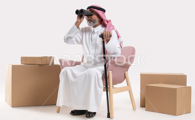 حب الاستكشاف والمغامرة, تحقيق الأهداف والطموحات المستقبلية, استخدام ناظور ثنائي العينين التلسكوب لرؤية الأشياء البعيدة, بورتريه لرجل كبير بالسن عربي خليجي سعودي يرتدي الثوب التقليدي والشماغ يمسك بيده العكاز وينظر إلى شيء ما عبر منظار الرؤية, خلفية بيضاء