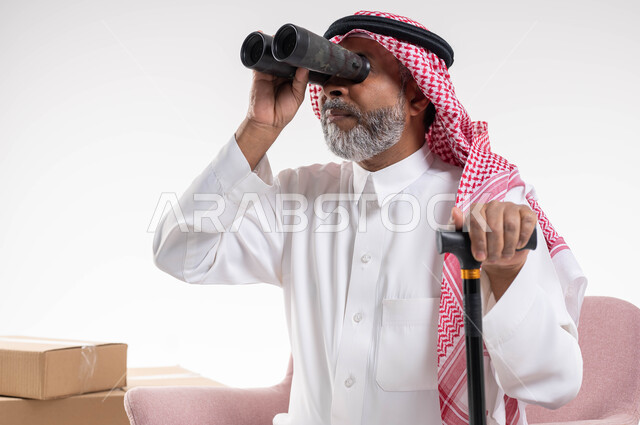 إيماءات التركيز والاستكشاف, بورتريه لرجل كبير بالسن عربي خليجي سعودي يرتدي الثوب والتقليدي والشماغ يمسك بيده العكاز وينظر إلى شيء ما عبر منظار التلسكوب ثنائي العينين, رؤية الأشياء البعيدة, خلفية بيضاء