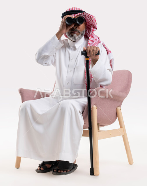 رؤية الأشياء البعيدة, الجلوس على الكرسي بإيماءات التركيز والاستكشاف, بورتريه لرجل كبير بالسن عربي خليجي سعودي يرتدي الثوب والتقليدي والشماغ يمسك بيده العكاز ويقوم بالنظر عبر منظار التلسكوب ثنائي العينين, خلفية بيضاء